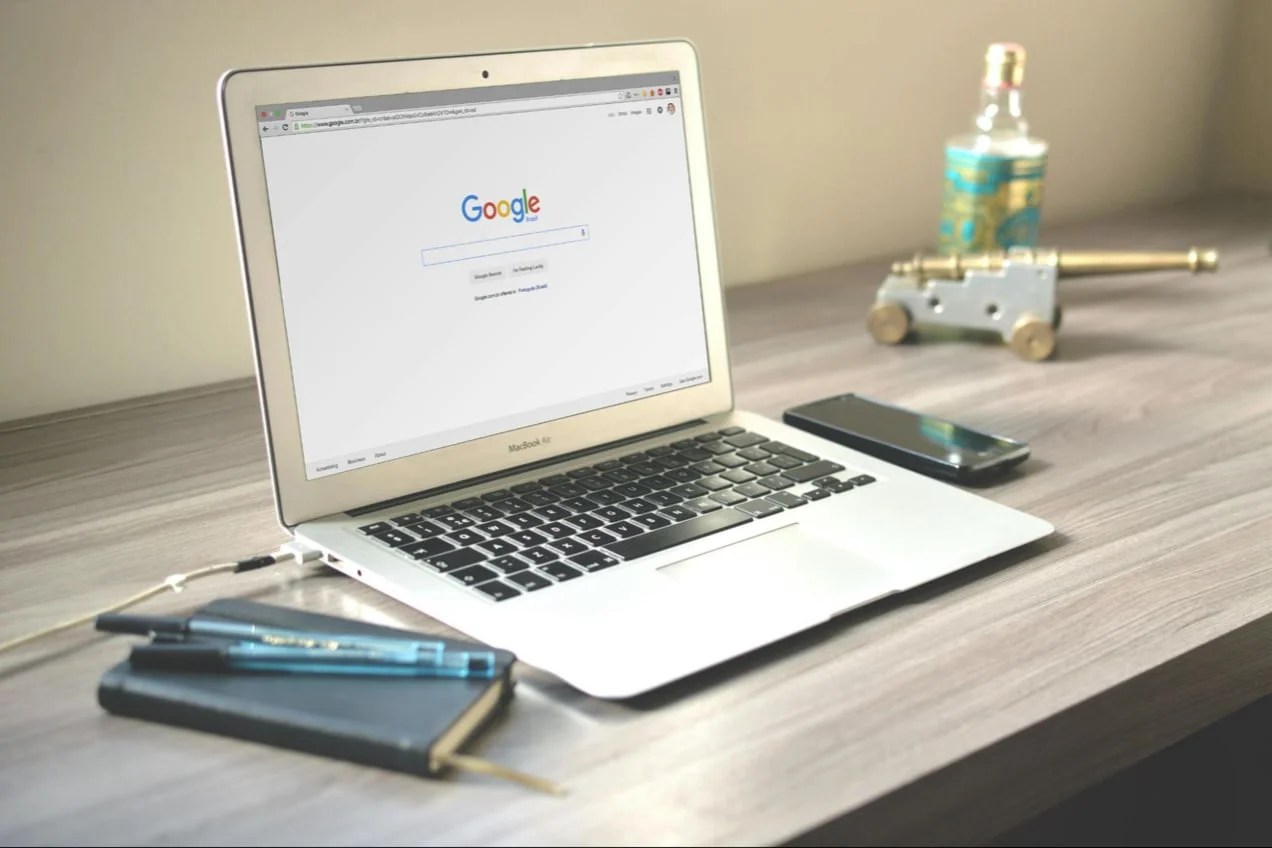 An apple laptop on a desk in an office that has the google homepage pulled up and ready for use.